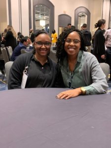 Two Black women smiling and sitting at a table during a conference.